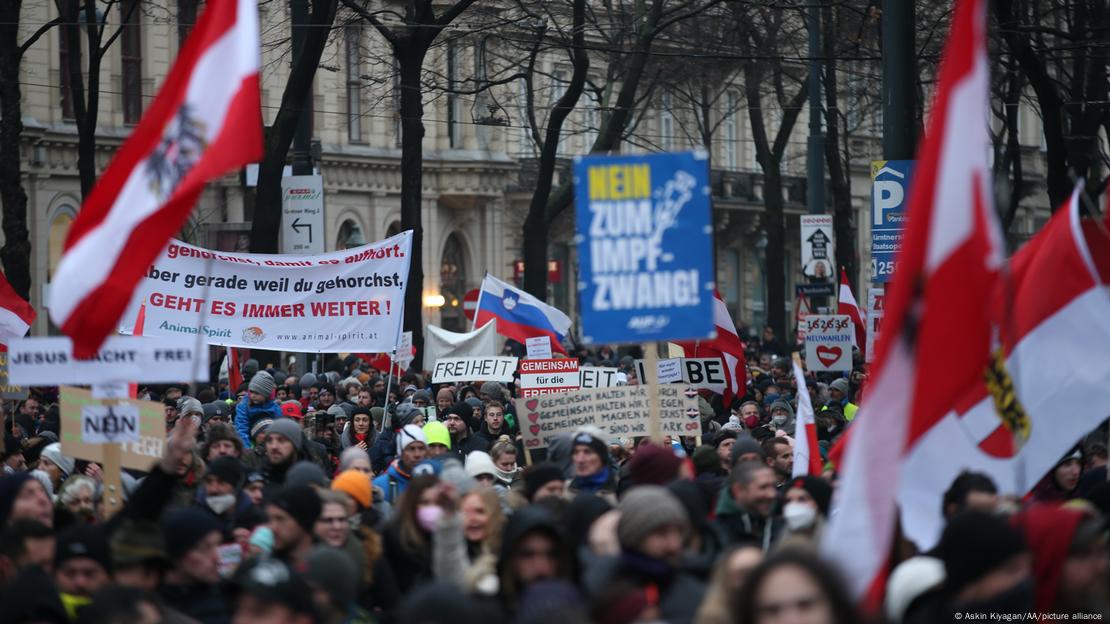 A imagem mostra centenas de pessoas aglomeradas durante um protesto contra a obrigatoriedade de vacinação contra o coronavírus, em Viena, Áustria. Elas portam bandeiras da Áustria e cartazes contrários à vacinação.