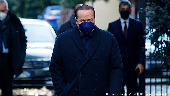 Silvio Berlusconi looking downward with his eyes closed and wearing a blue facemask