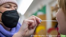 ARCHIV - 10.01.2022, Bayern, München: Eine Mutter entnimmt bei ihrem Kind einen Lollitest-Abstrich am Eingang zur Kita des Montessori-Zentrum. (zu dpa: «PCR-Tests in den Kitas: Der schwierige Weg zu mehr Sicherheit») Foto: Peter Kneffel/dpa +++ dpa-Bildfunk +++