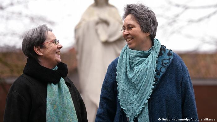 Zwei Frauen mittleren Alters, die sich gegenseitig Anlächeln, stehen vor einer Heiligenstatue