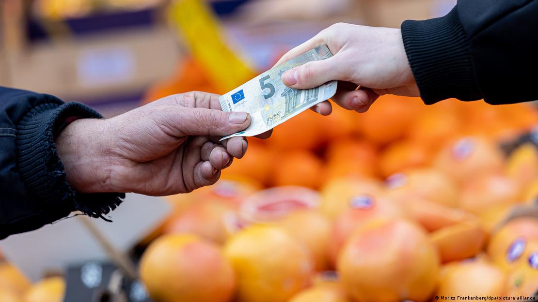 Dvije ruke drže novčanicu od 5 eura iznad štanda s voćem