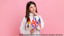 Thoughtful young girl holds flags in hands, looks away with pensive expression, studying languages, wearing white casual style sweater. Indoor studio shot isolated on pink background. || Modellfreigabe vorhanden