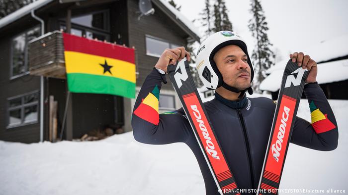 Carlos Maeder from Ghana carries his skis near a mountain hut draped with a Ghanaian flag 