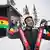 Carlos Mäder aus Ghana steht im Rennanzug auf seine Ski gestützt vor einer Berghütte an deren Balkon die Flagge Ghanas hängt