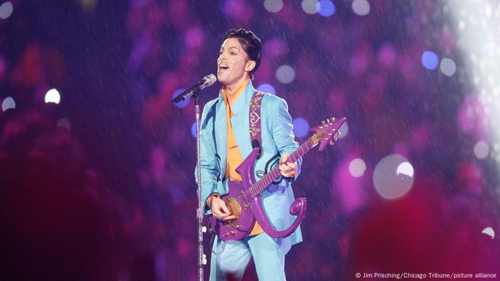 Prince, man in a light blue suit with a purple guitar stands in front of a microphone, singing in the rain