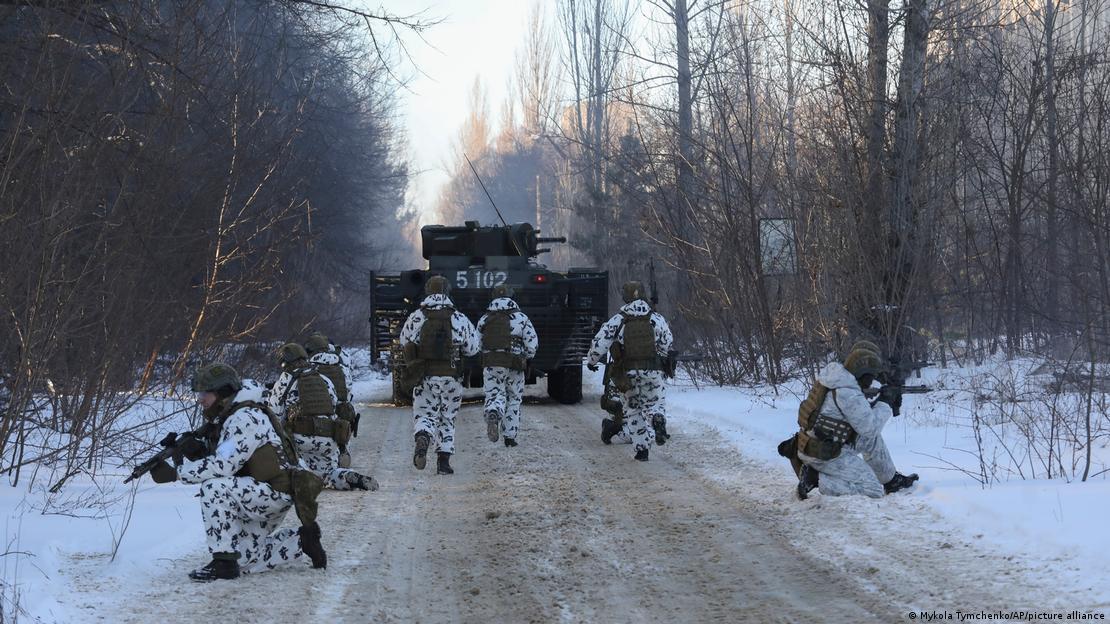 Soldados armados e vestindo roupas brancas com manchas pretas treinam em uma estrada com neve. Há um tanque de guerra na foto, 
