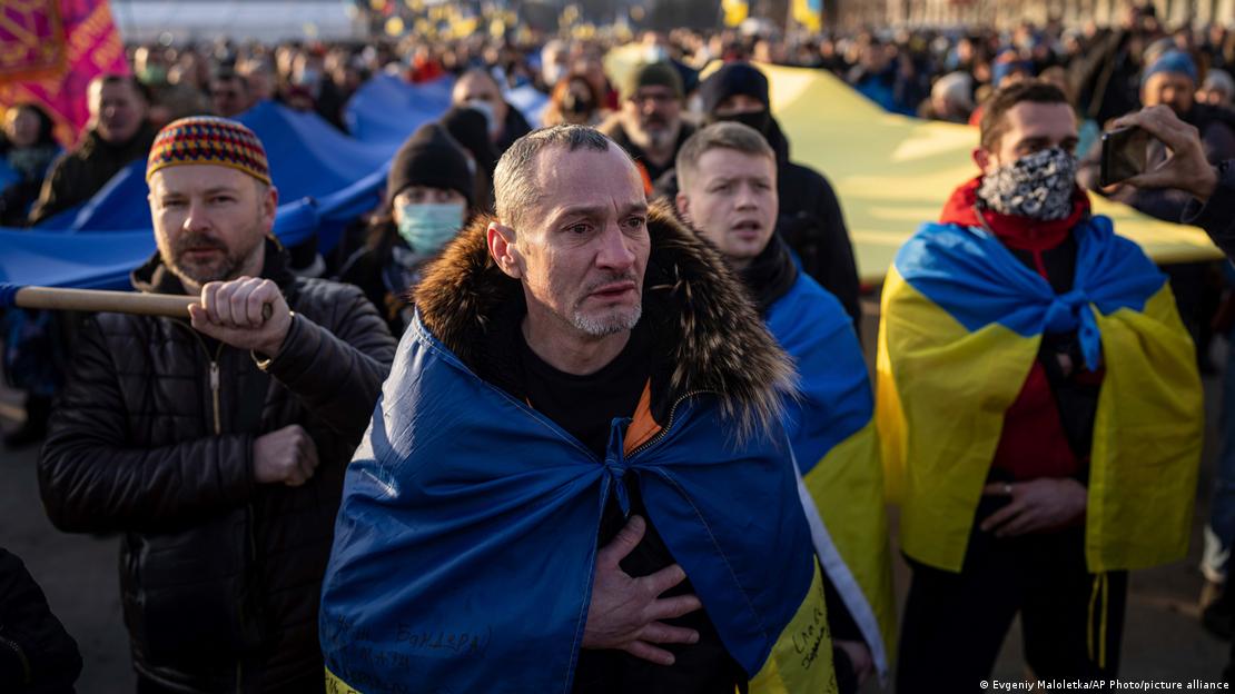 Um homem chora ao ouvir o hino nacional ucraniano durante protesto que ocorreu no sábado, 5 de fevereiro de 2022, na cidade de Kharkiv, a segunda maior da Ucrânia, a apenas cerca de 40 quilômetros de onde milhares de tropas russas estariam estacionadas, prontas para uma ofensiva. O homem tem uma expressão aflita. Ele está enrolado na bandeira da Ucrânia, e o mesmo ocorre com outros manifestantes que estão próximos.