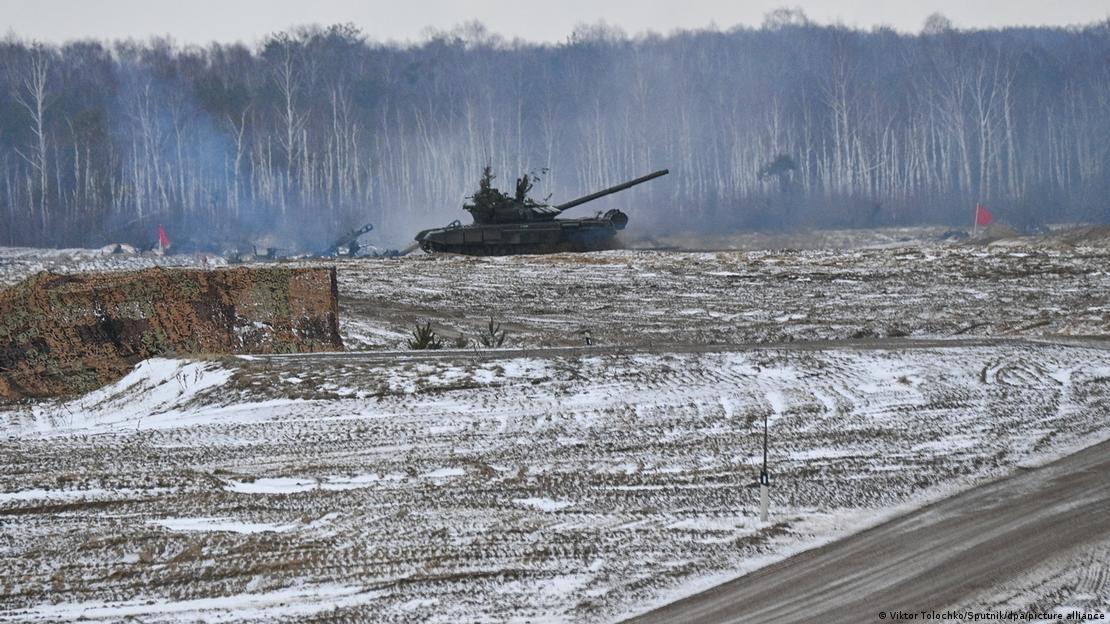 Um tanque de guerra trafega sobre uma área de treinamento militar na região de Brest, em Belarus. O tanque está ao fundo da imagem, em frente uma floresta. Um terreno com barro e neve e uma estrada à direita compõem a foto.