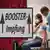Sign with 'Booster' in the foreground whilst someone in the background receives a vaccine jab