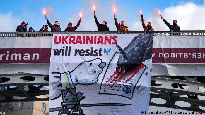 People stand on a bridge holding flares behind a banner that reads: Ukrainians will resist
