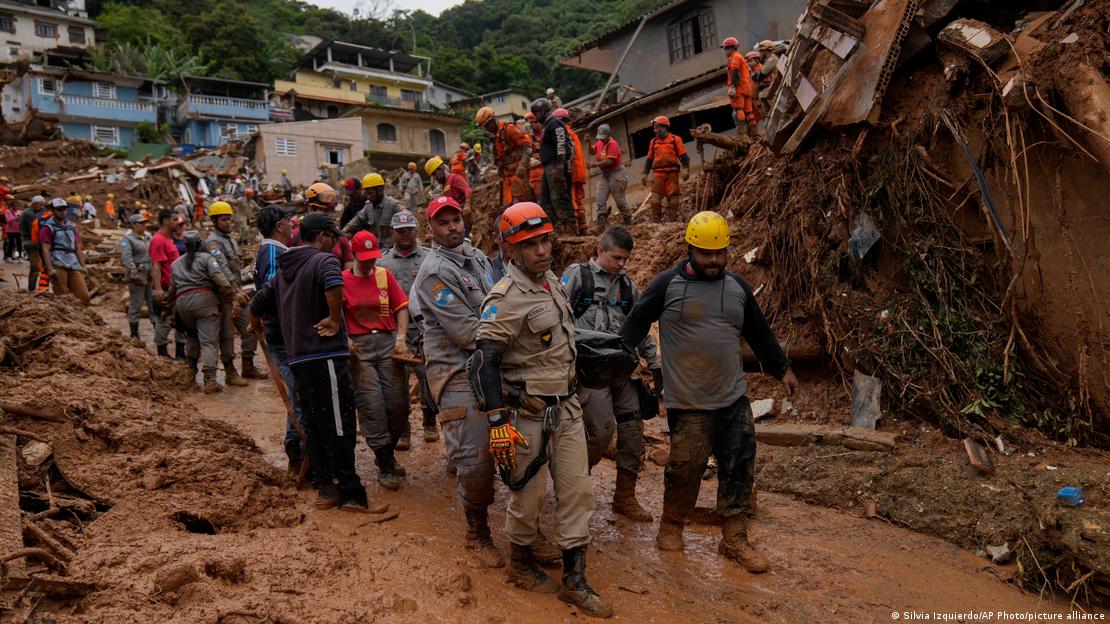 Bombeiros das equipes de resgate carregam mais um corpo encontrado em Petrópolis. Ao fundo da foto, há casas ao pé de um morro.