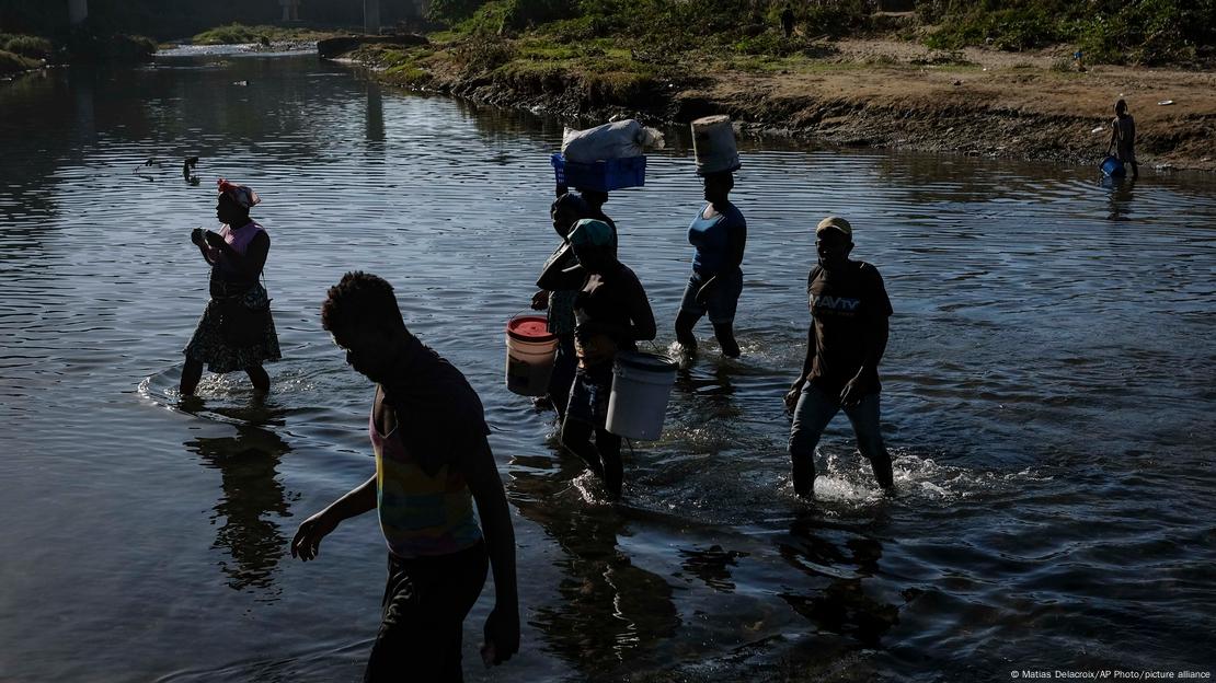 Pessoas passam a pé por um rio, Algumas carregam baldes. 