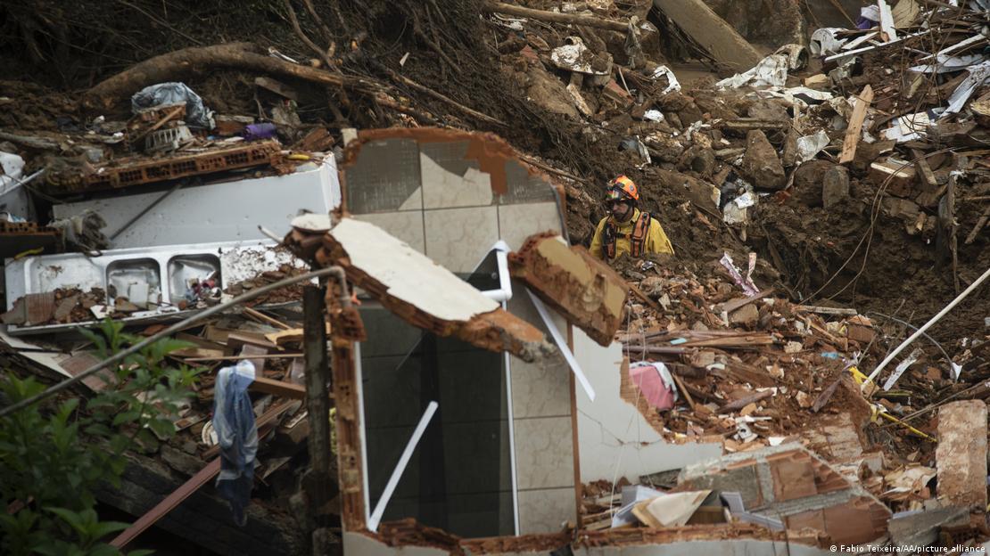 A imagem mostra destroços de casas que foram levadas por enxuradas e deslizamentos. Um bombeiro, que veste roupa laranja e um capacete, está ao fundo da foto, em meio a uma área cheia de terra e pedras.