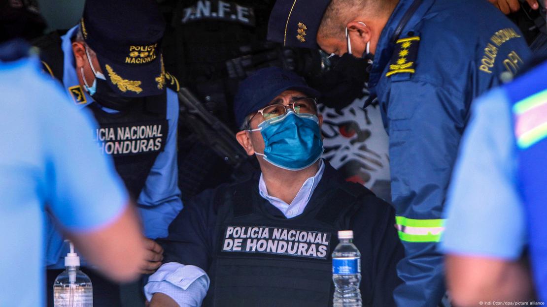 O ex-presidente de Honduras, Juan Orlando Hernández, veste um colete à prova de balas preto, da Polícia Nacional, sobre uma camisa clara. Ele está sentado e conversa com policiais. Veste boné, óculos e máscara. Em pé, dois policiais o cercam.