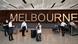 A imagem mostra sete pessoas com malas chegando ao Aeroporto Internacional de Melbourne, na Austrália. Ao fundo, há um grande letreiro escrito Melbourne. À frente, ao centro, uma mulher carrega duas malas. Ela veste máscara e uma blusa clara.