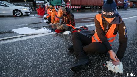 Einige Personen mit orangefarbenen Westen sitzen auf einer Fahrbahn und kleben sich selbst daran fest.