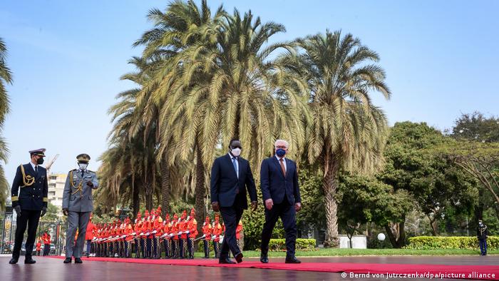 Senegalese President Macky Sall (l) and German President Frank-Walter Steinmeier (r) walk along a red carpet as a military band plays in the background under blue skies and palm trees