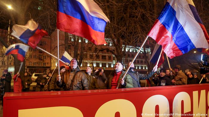 Hinter einen großen Schild mit der Aufschrift Rossia - Russland schwenken Männer mehrere russische Nationalflaggen