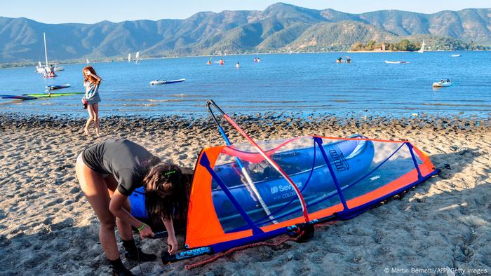 En una playa de arena junto a una laguna, una mujer fija una vela a una tabla de surf, al fondo se ve un niño de pie.