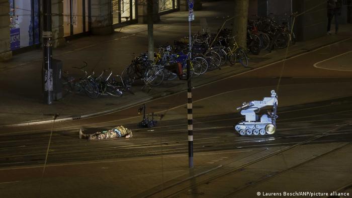 A man in camouflage clothing is lying next to a police robot on the street