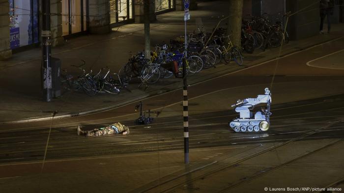 A man in camouflage clothing is lying next to a police robot on the street