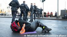 23.02.2022, Eine Klimaaktivistin der Aktion „Aufstand der letzten Generation“ hat sich auf einer Straße am Flughafen festgeklebt. Hinter stehen Polizeibeamte.