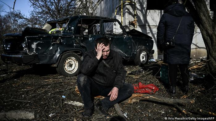 Мужчина сидит перед разбомбленным автомобилем у жилого дома в Чугуево.