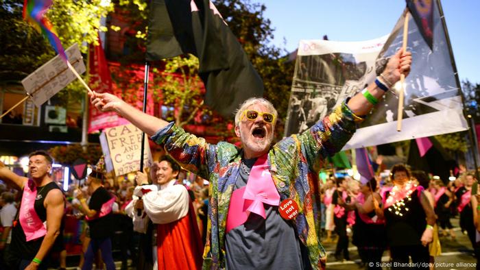 An exuberant man waves a rainbow flag and placard at a Sydney pride parade, scores of revelers are seen in the background