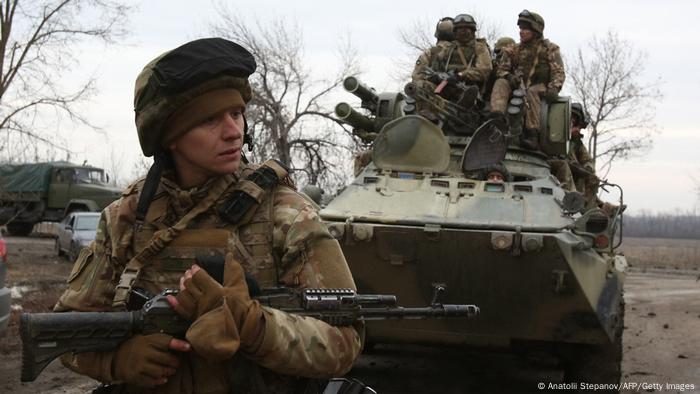 A soldier holding a gun stands in front of a tank