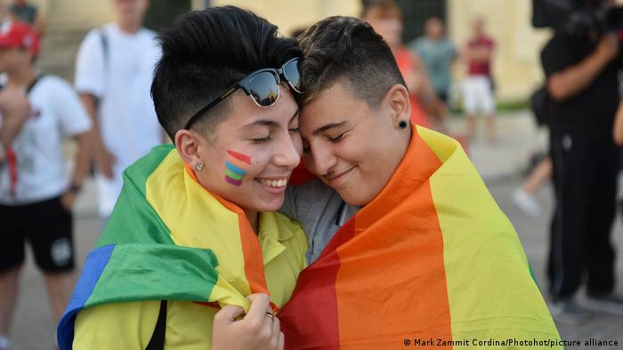 A couple wearing a rainbow flag over their shoulders embrace