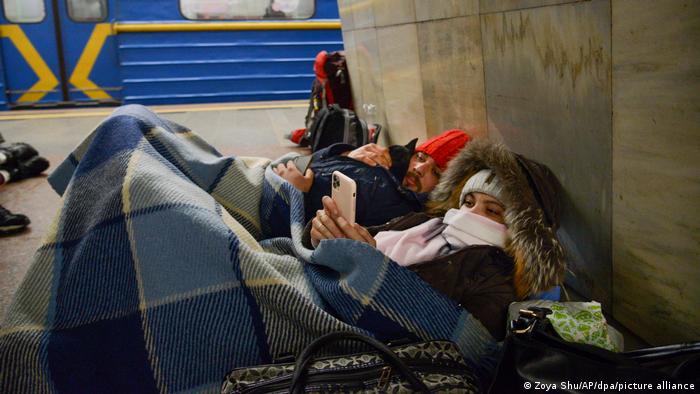 Two persons lie in a subway station, covered in blankets