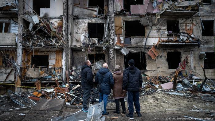 Four people, seen from behind, stand in front of a destroyed apartment building in Kyiv