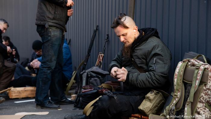 A Ukrainian man with a trendy haircut in civilian clothes, sitting on the ground in front of a wall beside a patterned backpack, loading a rifle. Other men in civilian clothes and rifles in the background.