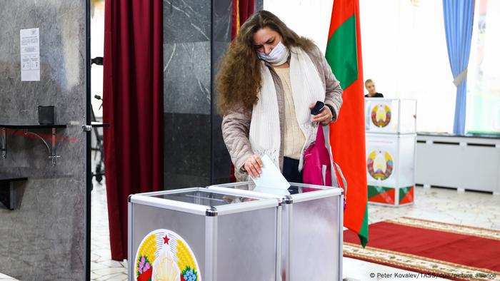 A woman diposits a ballot into an urn with the Belarusian coat of arms on it