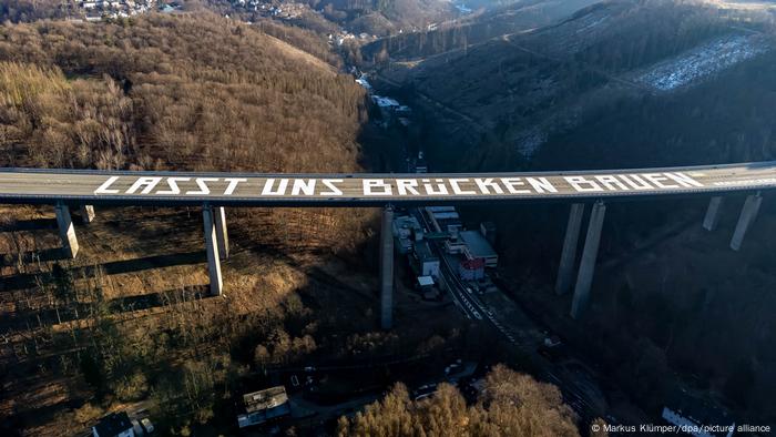The words Lasst uns Brücken Bauen appear on a tall highway overpass spanning a valley between wooded hills.