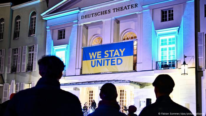  Über dem Eingang des Deutschen Theaters hängt ein blau-gelbes Banner mit der Aufschrift We stay united . Davor stehen Menschen 