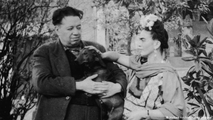 Eine Schwarz-Weiß-Aufnahme zeigt Diego Rivera und Frida Kahlo, die eine Hand auf den Kopf eines Hundes legt, den Rivera im Arm hält.