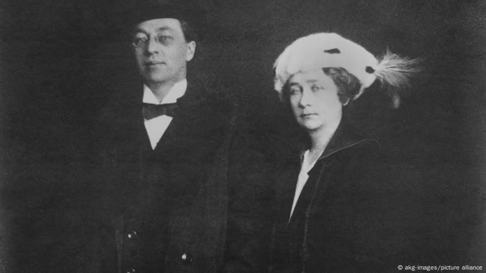 Wassily Kandinsky und Gabriele Münter stehen in einer Schwarz-weiß-Aufnahme nebeneinander, er trägt Anzug und Fliege, sie einen weißen Hut mit Feder.