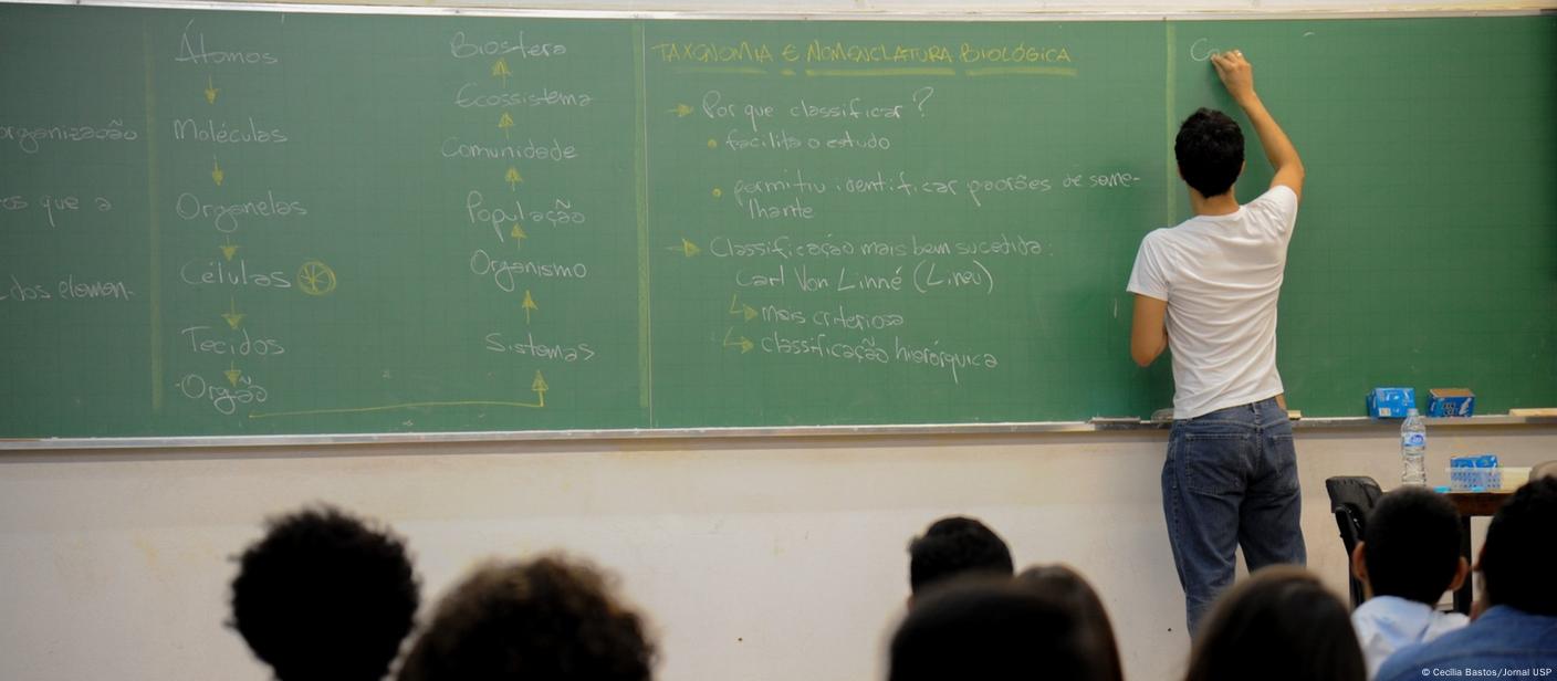 Professor escreve em quadro em uma sala de aula. Fotografia mostra alunos de costas para a câmera e olhando para a lousa.