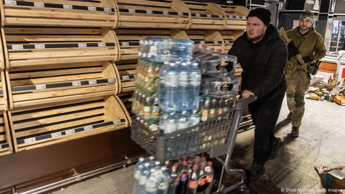 Two men walk down an aisle with empty shelves, pushing a cart stacked with water and soft drink bottles 