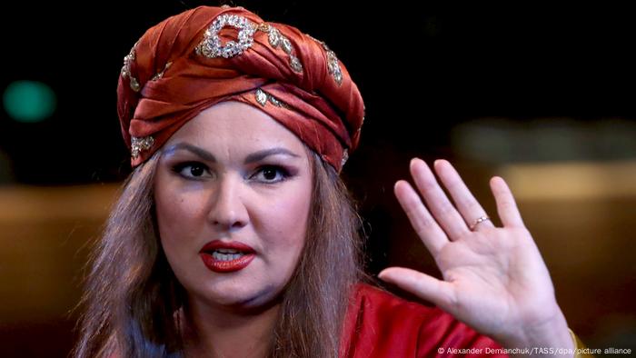 Anna Netrebko hält ihre linke Hand ins Bild, auf dem Kopf trägt sie einen Turban aus elegantem Stoff
