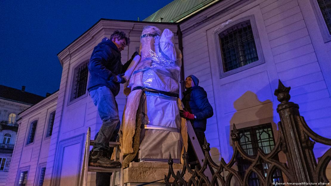Foto à noite mostra dois homens embrulhando uma escultura grande em frente a um prédio