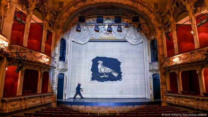 A stage curtain bearing a blue and white dove hangs in an empty theater as a man walks across the stage.