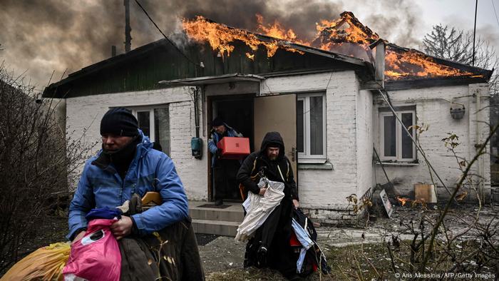 Single storey white house with a burning roof, three people carry items out 