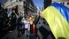Foto mostra um protesto, com várias pessoas. Bem à frente está uma mulher, com flores na cabeça, segurando uma grande bandeira da Ucrânia. 