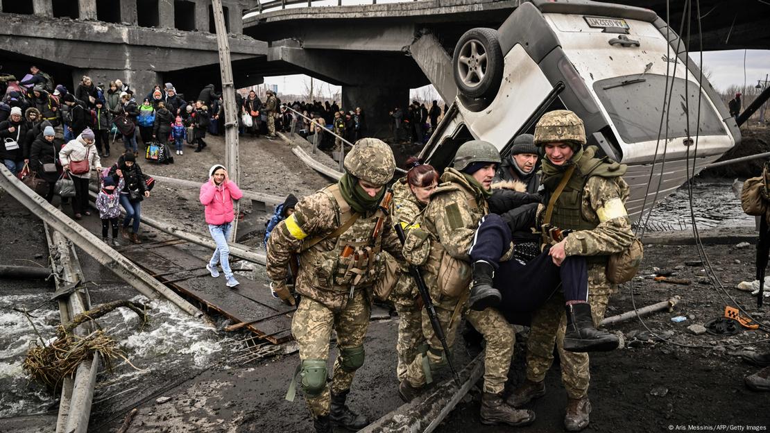 Foto mostra soldados russos carregando uma pessoa. Ao fundo, uma fila de pessoas passa por uma ponte improvisada.