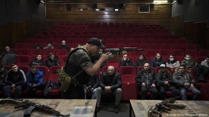 Men sit on red chairs in a movie theater, watching one man at the front with a rifle 