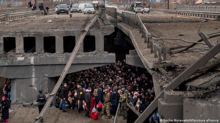 A large crowd underneath a destroyed bridge, cars still on the road 