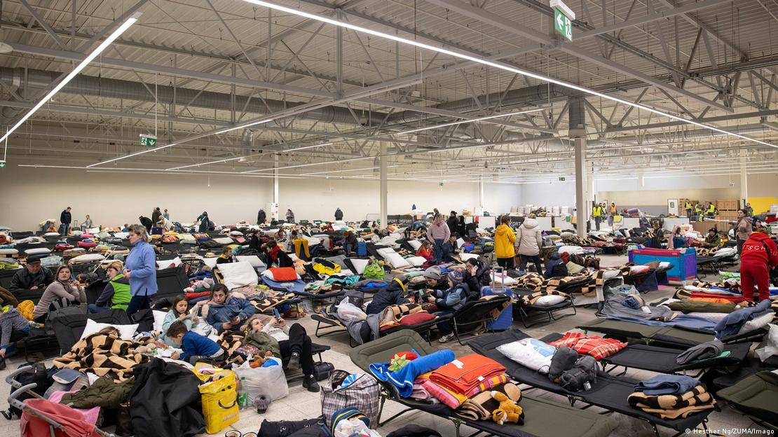 A imagem mostra diversos refugiados ucranianos em um centro de refugiados na Polônia. Há muitas camas, cobertores, travesseiros, mochilas, malas e sacolas em uma vasta área coberta. Algumas pessoas caminham pelo local.