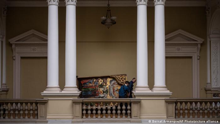 In einem neoklassischen Gebäude mit hohen weißen Säulen sieht man einige Menschen, die ein altes, goldgerahmtes, mannshohes Gemälde tragen